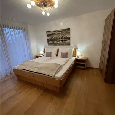 Ein komfortables Schlafzimmer mit einem großen Doppelbett und warmen Holzfußboden. Sanfte Beleuchtung und Vorhänge sorgen für eine gemütliche Atmosphäre.