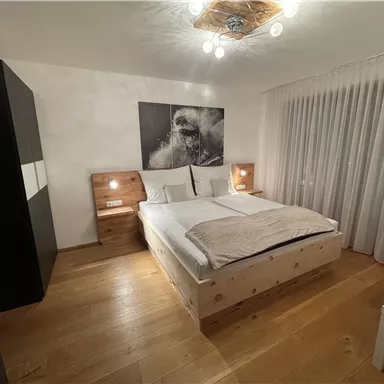 Ein modernes Schlafzimmer mit einem großen Bett und Holzboden. Die Wände sind hell und es gibt große Fenster mit Vorhängen.