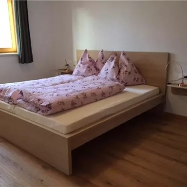 Ein einfaches Schlafzimmer mit einem Holzbett und rosa Bettwäsche. Das Zimmer hat ein Fenster mit Vorhängen und einen kleinen Nachttisch.