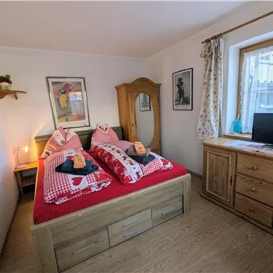 Ein gemütliches Schlafzimmer mit zwei Einzelbetten, roten Bettdecken und dekorativen Kissen. An der Wand hängen ein paar Bilder, und ein Holzschrank steht neben einem Fenster.