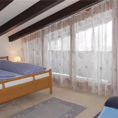 Ein gemütliches Schlafzimmer mit einem Doppelbett und blauen Bettdecken. Große Fenster mit Vorhängen sorgen für viel natürliches Licht.