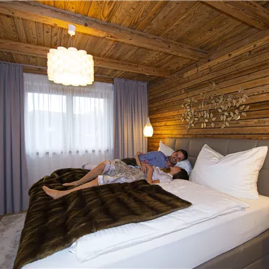 A cozy bedroom with wooden paneling and modern design. A couple is relaxed in the large bed under a warm blanket.
