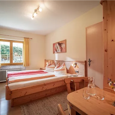 A bright room with two beds and a small table. In front of the window are curtains and a floral decoration on the table.