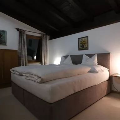 Ein gemütliches Schlafzimmer mit einem großen Bett und hellen Wänden. Die Holzdecke und die sanfte Beleuchtung sorgen für eine warme Atmosphäre.