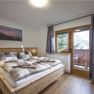 A cozy bedroom with a large bed and wooden details. There is a balcony overlooking nature and beautiful curtains.