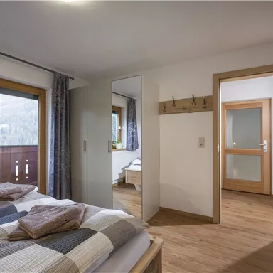 A bright bedroom with a large bed and a mirror. The windows offer a view outside, and the room has a modern, cozy style.