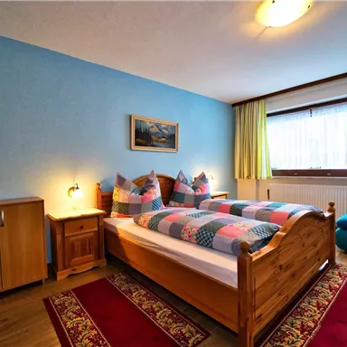 Ein gemütliches Schlafzimmer mit zwei Betten, bunten Kissen und einem blauen Wanddesign. Eine Ecke mit einem Sessel und Holzschränken sorgt für eine einladende Atmosphäre.