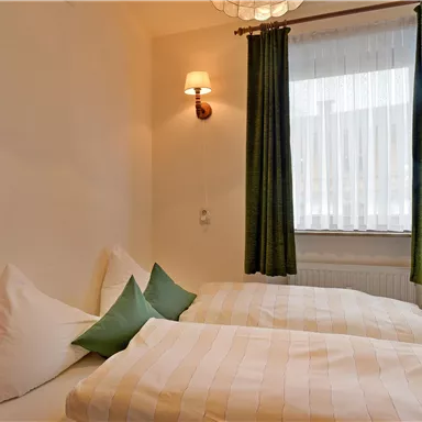 Das Bild zeigt ein gemütliches Schlafzimmer mit zwei Betten und grünen Kissen. Vor dem Fenster hängen helle Vorhänge, die Tageslicht hereinlassen.