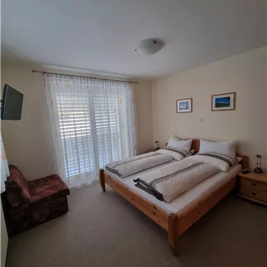 Ein helles Schlafzimmer mit zwei Einzelbetten und einem gemütlichen Sessel. Große Fenster mit Lichtvorhängen sorgen für eine angenehme Atmosphäre.