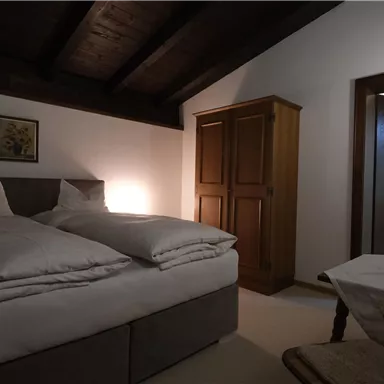 Ein gemütliches Zimmer mit einem Doppelbett und Holzmöbeln. Es gibt einen Stuhl und einen Tisch, sowie eine Tür, die zu einem Badezimmer führt.