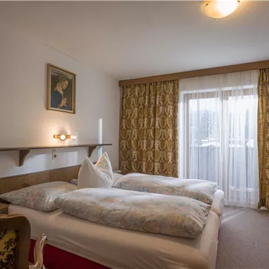 A cozy bedroom with two beds and a warm color scheme. Large windows provide daylight and a view outside.