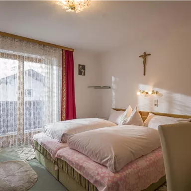 A cozy bedroom with two beds and soft colors. Large windows with curtains provide plenty of daylight.