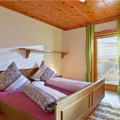 A cozy bedroom with two beds and decorative pillows. Large windows let in plenty of daylight and show green curtains.