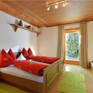 A cozy bedroom with a double bed and red pillows. Large windows let in plenty of light and offer a view of the outdoors.