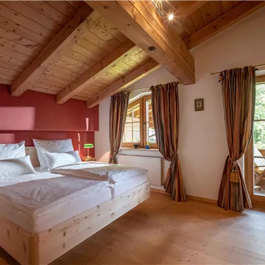 A cozy bedroom with a wooden beam ceiling and a large window. The room is bright and inviting, featuring a double bed and warm colors.