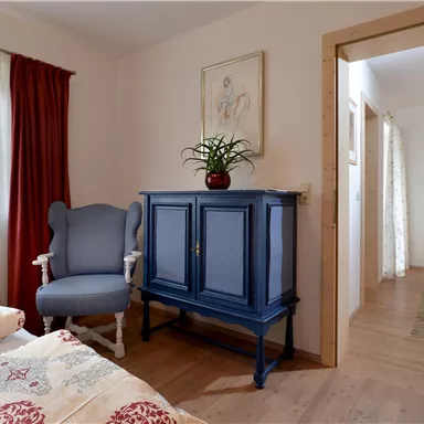 A cozy bedroom with inviting colors and a blue wardrobe. In the background, a bright room with a seating area is visible.