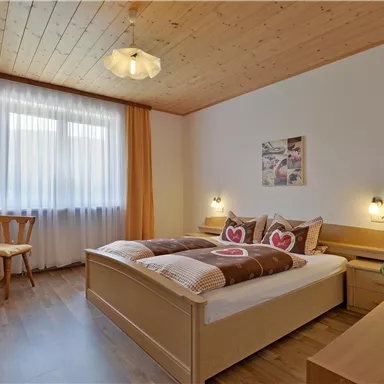Ein gemütliches Schlafzimmer mit einem Doppelbett und Dekokissen in Herzform. Der Raum hat große Fenster, helles Holz und eine freundliche Atmosphäre.