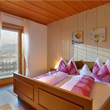 A cozy bedroom with a double bed and purple pillows. Next to it a window with plenty of daylight and a view outside.