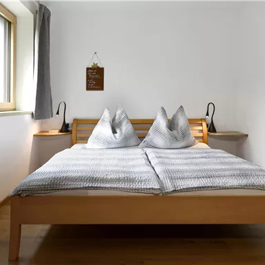 Ein helles Schlafzimmer mit einem Doppelbett und zwei Kopfkissen. Es gibt Fenster, die Licht hereinzulassen, sowie eine gemütliche Holzdekoration.