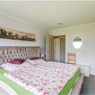 A cozy bedroom with a large upholstered bed and colorful bedcovers. The walls are light, and there is a round mirror as well as a picture of mountains.