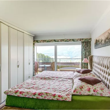 A cozy bedroom with a double bed and colorful bedding. Large windows offer a view of nature and plenty of daylight.