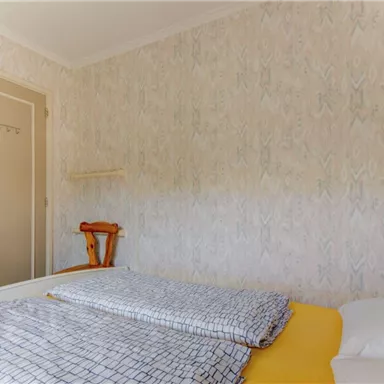 A simple bedroom with a bed and a yellow duvet. The walls are decorated with a light, patterned wallpaper, and a door leads outside.