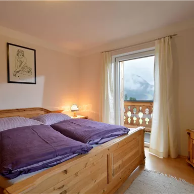 Ein gemütliches Schlafzimmer mit einem Holzbett und lila Kissen. Große Fenster bieten einen Blick auf die Berge.