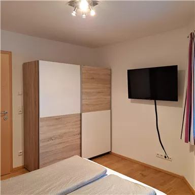 Ein modernes Schlafzimmer mit einem Doppelbett, einem Schrank und einem Fernseher an der Wand. Das Zimmer hat eine helle und gemütliche Atmosphäre.