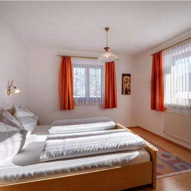 A bright bedroom with a double bed and cozy decoration. Large windows provide plenty of daylight and the room has orange curtains.