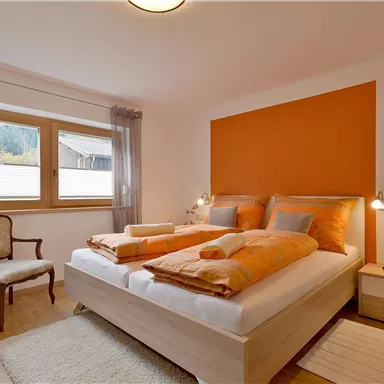 A cozy bedroom with a large bed and orange accents. Large windows provide plenty of daylight and a pleasant atmosphere.