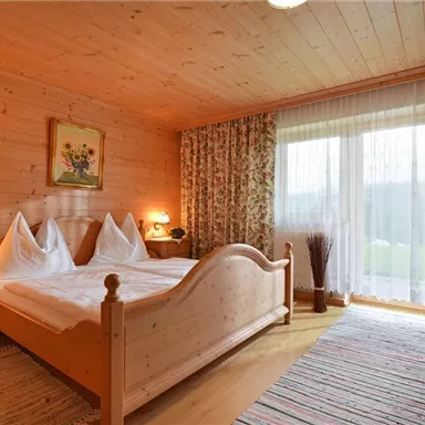 A cozy bedroom with wood paneling and a large window. There is a double bed and pretty floral curtains.