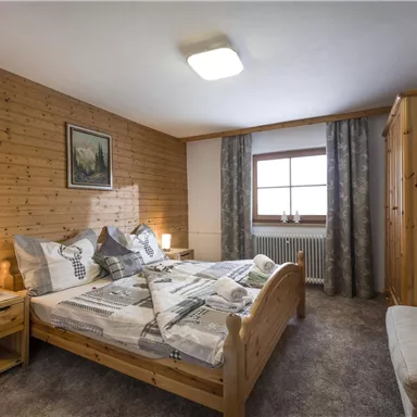 A cozy bedroom with wood paneling and a large window. The room is furnished with a double bed, nightstands, and a comfortable sofa.
