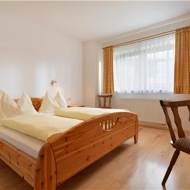A cozy bedroom with a double bed and two pillows. The room has large windows and warm wooden details.