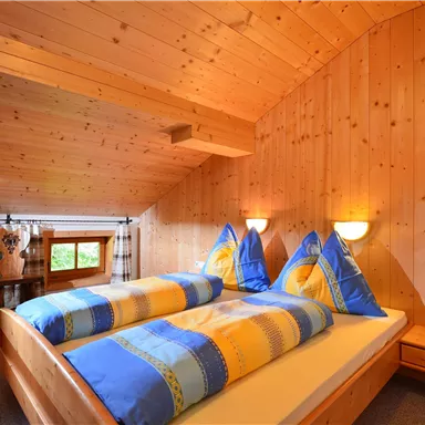 Ein gemütliches Zimmer mit Holzverkleidung und einem großen Doppelbett. Die bunten Kopfkissen und warmes Licht sorgen für eine einladende Atmosphäre.