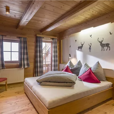 A cozy bedroom with wooden paneling and a large bed. The wall features deer motifs and there is a window front with curtains.