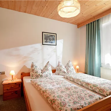 Ein gemütliches Schlafzimmer mit zwei Betten, hellen Holzmöbeln und einem Fenster mit Vorhängen. An der Wand hängt ein Bild, und es gibt sanfte Beleuchtung mit Nachttischlampen.