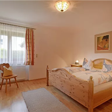 Ein gemütliches Schlafzimmer mit einem Doppelbett und Holzmöbeln. Das Zimmer hat große Fenster, Vorhänge in warmen Farben und einen Teppich auf dem Boden.