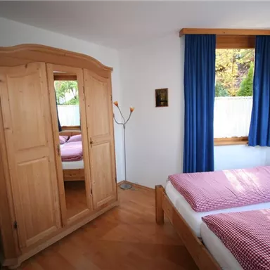 A cozy bedroom with two beds and a large wooden wardrobe. The windows are adorned with blue curtains and let in plenty of light.