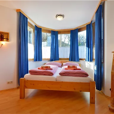 A cozy bedroom with a double bed and colorful bedding. Large windows with blue curtains let in a lot of light.