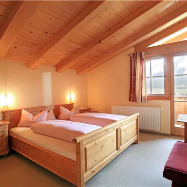 A cozy bedroom with wooden furniture and a large window. The atmosphere is bright and welcoming.