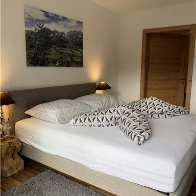 A cozy bedroom with a large bed and stylish cushions. The walls are painted in neutral tones, and a picture with a landscape motif hangs above the bed.