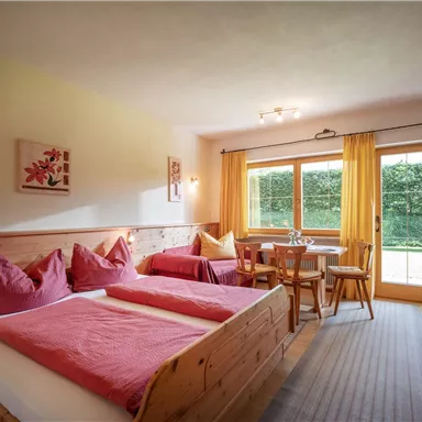 A cozy room with a double bed and pink pillows. There is a dining table and large windows overlooking a well-kept garden.