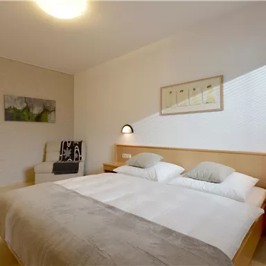 Ein modernes Schlafzimmer mit zwei Einzelbetten, einem bequemen Sessel und sanfter Beleuchtung. Die Wände sind hell und minimalistisch dekoriert.