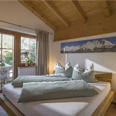 A cozy bedroom with a large window and wooden details. The beds are decorated with green pillows and blankets.