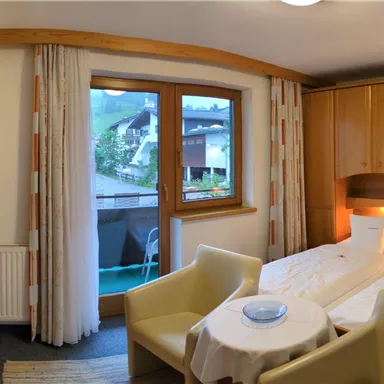 Ein helles Zimmer mit Holzmöbeln und einem großen Fenster. Zwei Betten und ein Tisch mit Stühlen bieten gemütlichen Platz.