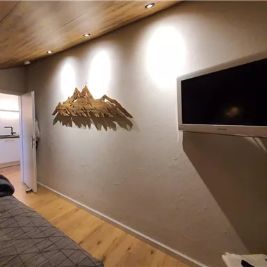 A cozy room with a TV on the wall and a decorative wooden sculpture. In the background, an open kitchen is visible.