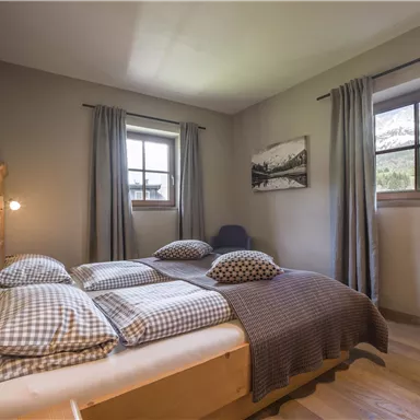 Ein gemütliches Schlafzimmer mit einem großen Bett und karierten Kissen. Fenster bieten einen Blick auf die Natur, während Vorhänge das Licht sanft filtern.