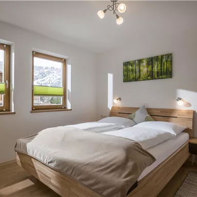 Ein gemütliches Schlafzimmer mit einem großen Bett und zwei Fenstern. Die Wände sind hell gestrichen und ein grünes Bild verleiht dem Raum eine frische Note.