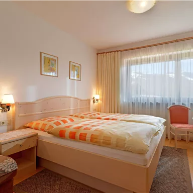 Ein gemütliches Schlafzimmer mit einem großen Doppelbett und hellen Vorhängen. Ein kleiner Tisch und Stühle ergänzen den Raum, der freundlich und einladend wirkt.