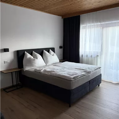 Ein gemütliches Schlafzimmer mit einem Doppelbett und zwei Kissen. Große Fenster lassen viel Licht herein und der Boden ist aus Holz.